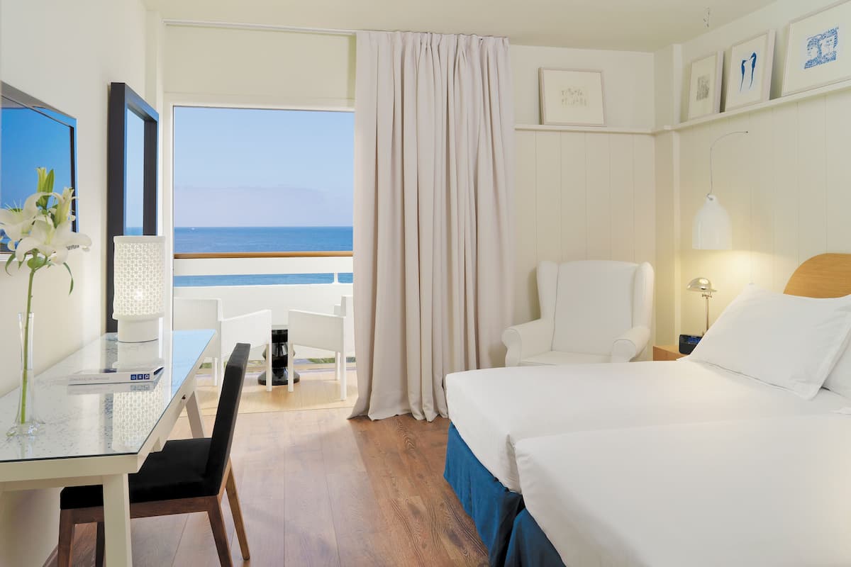 Una habitación de hotel con cama, escritorio con computadora, silla, lámpara y vista al océano.