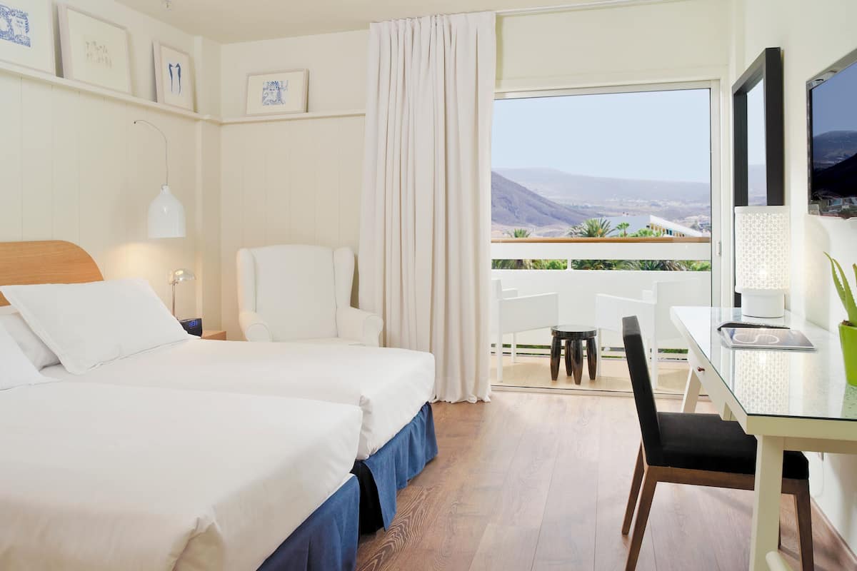 Habitación de hotel con dos camas, un escritorio con ordenador, una silla y un balcón con vistas.