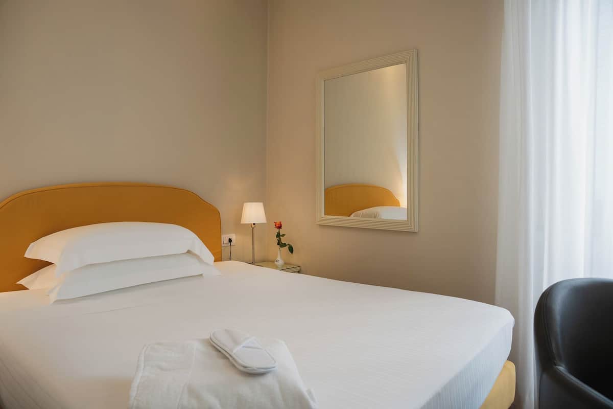 A hotel room with a bed, a nightstand, a lamp, a vase with a flower, and a chair.
