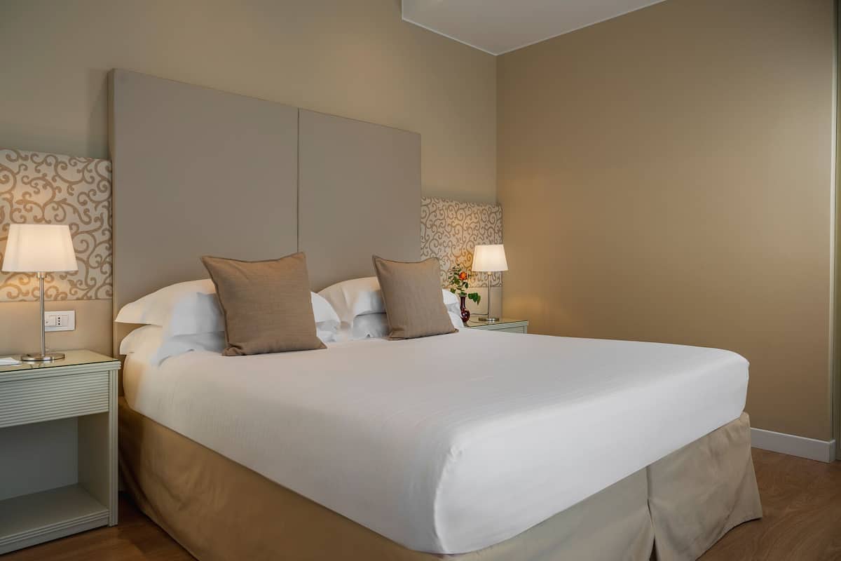 A hotel room with a large bed, two bedside tables with lamps, a nightstand, and a patterned wallpaper.