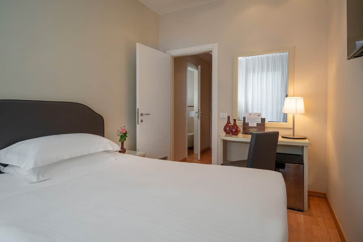 A hotel room with a bed, a desk, a chair, a lamp, and a mirror.