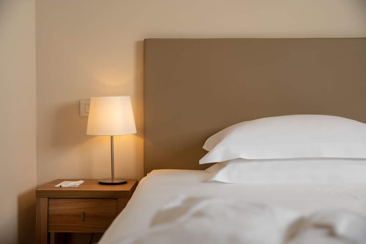 A neatly made bed with white linens and pillows, a bedside table with a lamp, and a wooden nightstand.