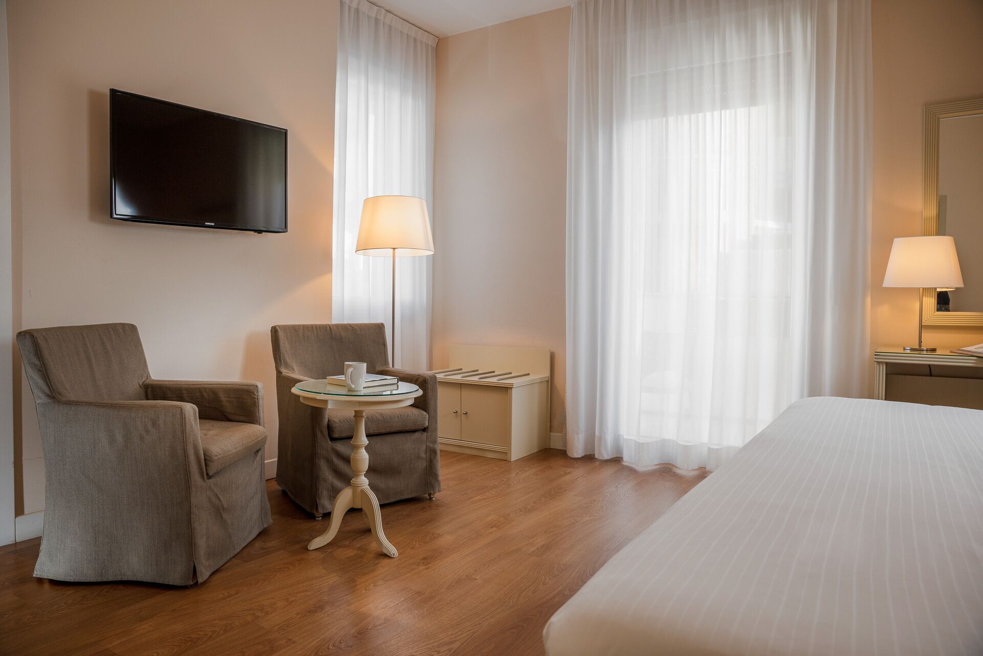 Hotelkamer met een bed, twee fauteuils, een bijzettafeltje met een lamp, een aan de muur bevestigde televisie en een groot raam met dunne gordijnen.