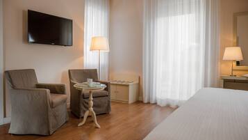 Hotelkamer met een bed, twee fauteuils, een bijzettafeltje met een lamp, een aan de muur bevestigde televisie en een groot raam met dunne gordijnen.