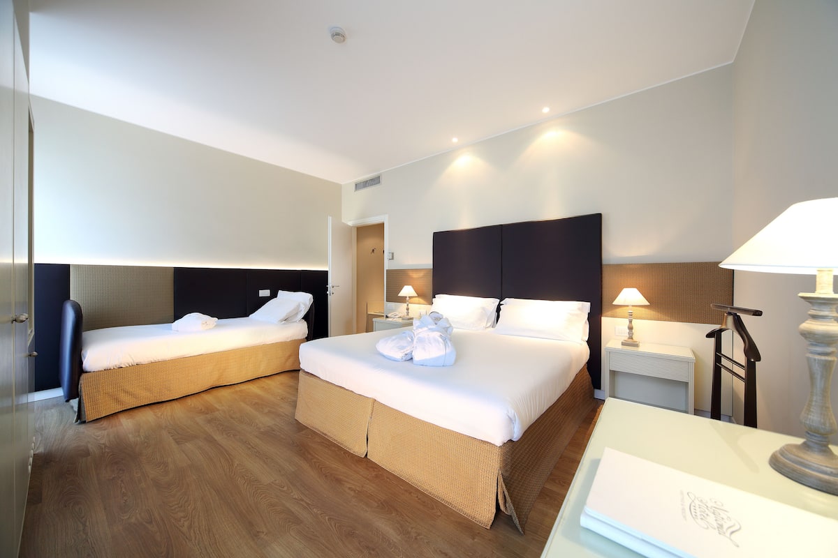 A hotel room with two single beds, a wooden floor, a nightstand with a lamp, and a bedside table with a lamp.
