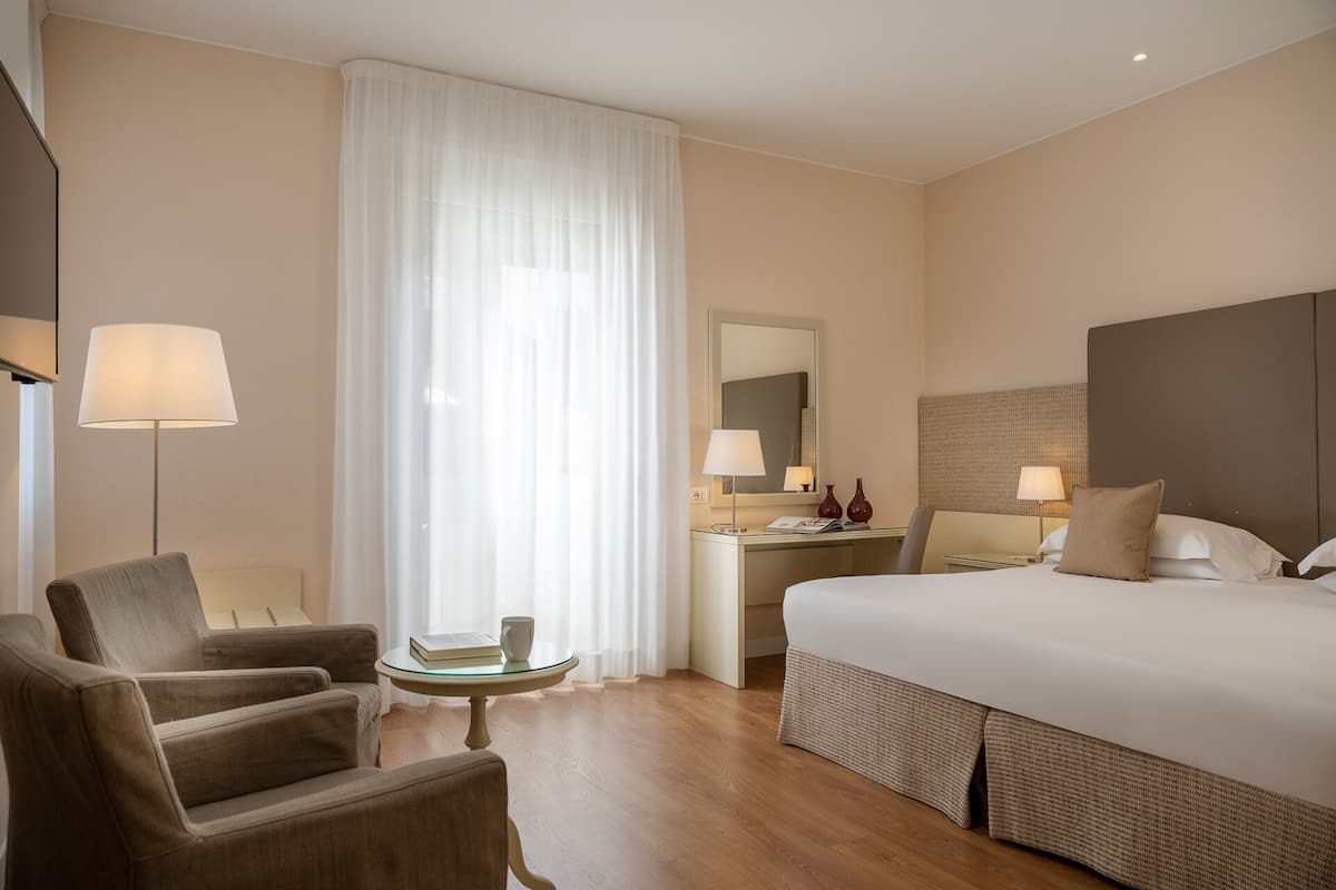 A hotel room with a bed, two armchairs, a small table, a desk with a lamp, and a large window with sheer curtains.