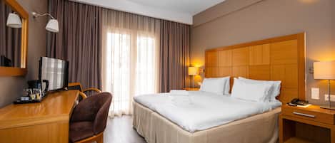 A hotel room with a bed, a desk with a coffee maker, a chair, and a nightstand with a lamp.