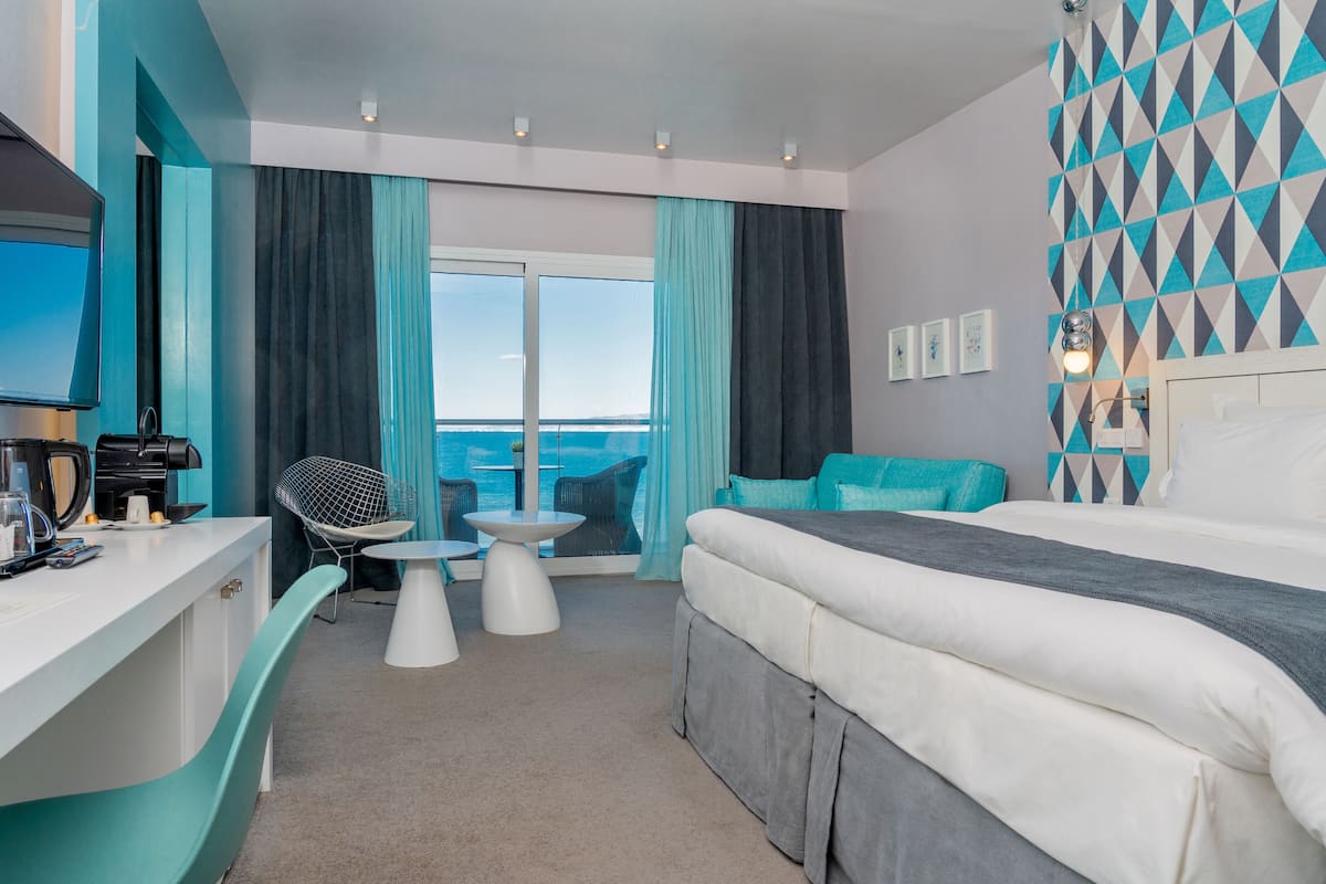 Habitación de hotel moderna con una cama grande, un escritorio con cafetera, una silla, una mesita y vistas al mar a través de amplios ventanales.