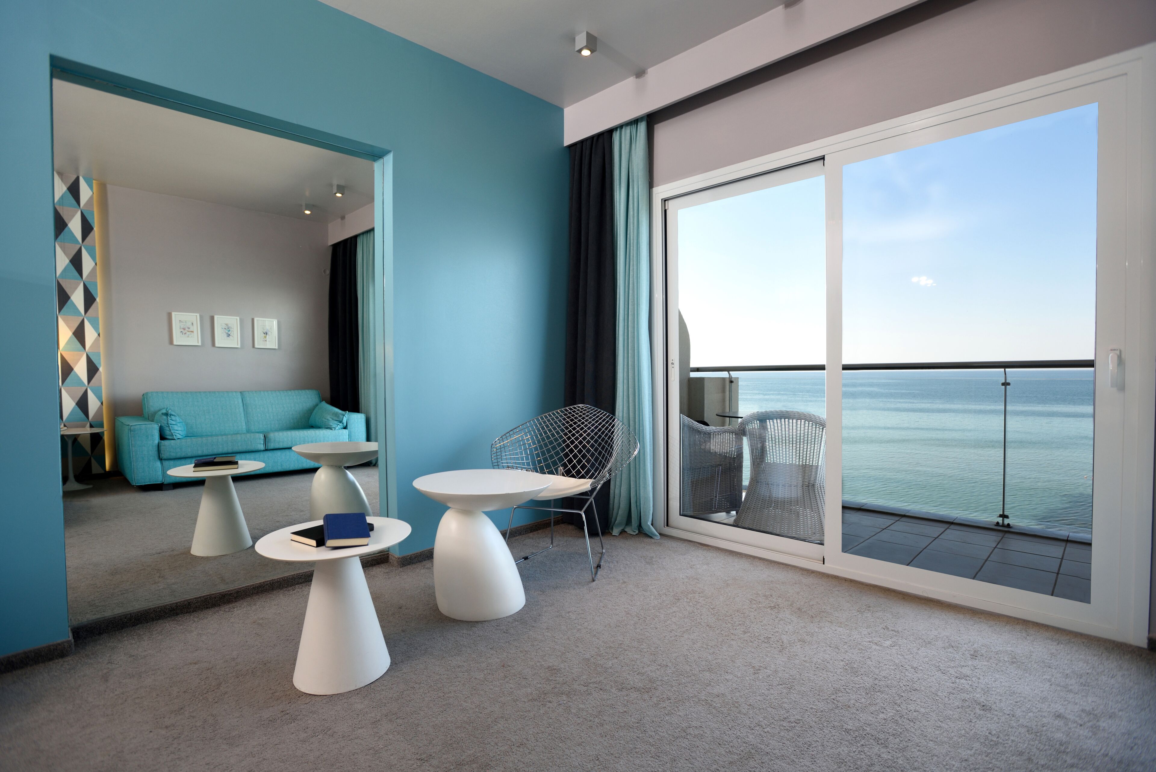 Una habitación moderna con una pared azul de acento, un gran ventanal corredero a un balcón, un sofá, dos mesas blancas y una silla de estructura metálica.