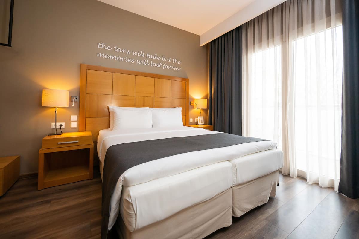Habitación de hotel moderna con una cama grande, dos mesitas de noche, un cabecero con lámpara y una ventana con cortinas.