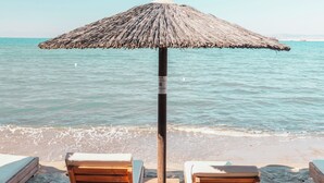Dos sillones de madera bajo un toldo de paja en una playa arenosa, con el océano al fondo.