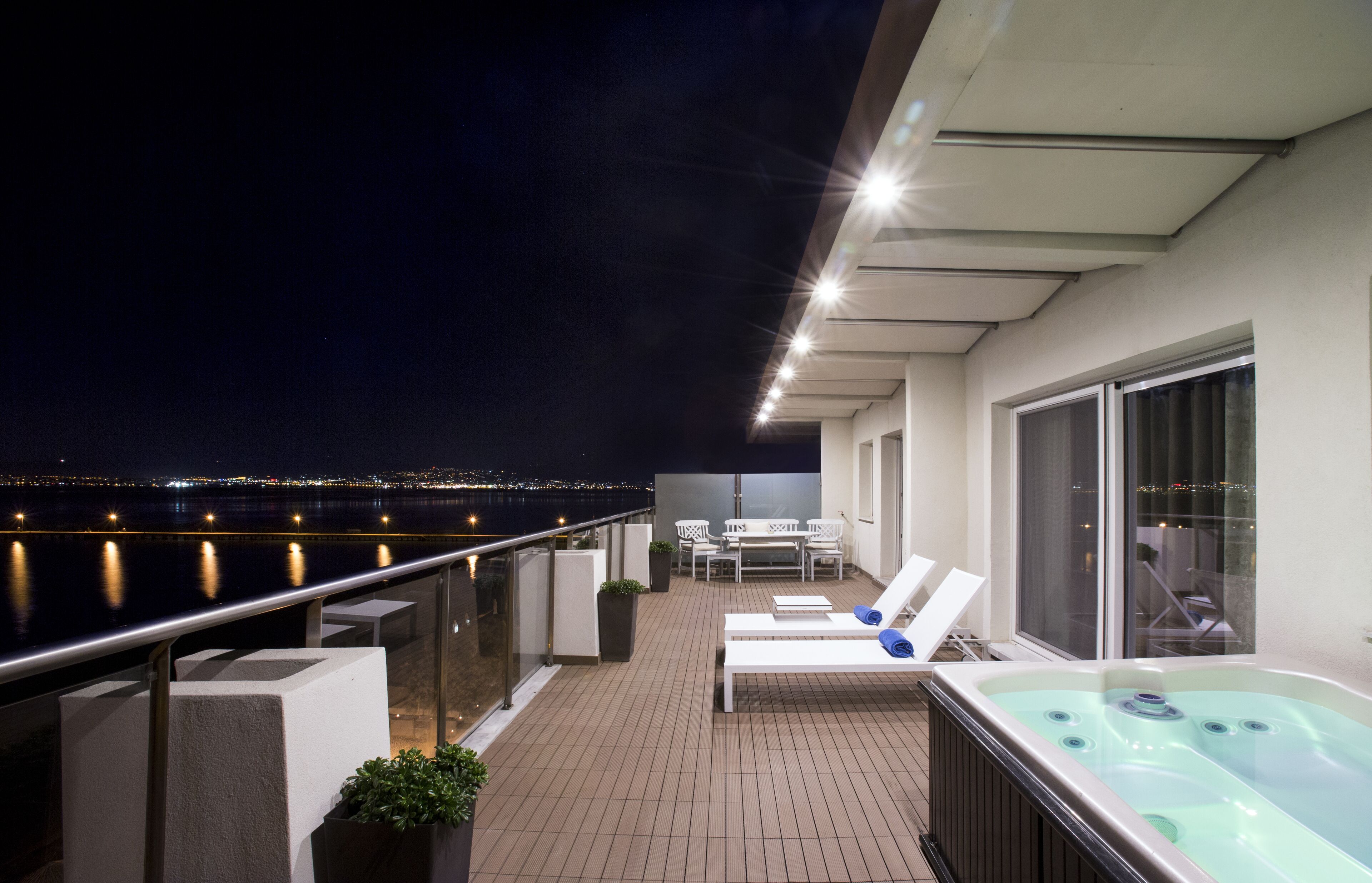Terraza en la azotea con jacuzzi, sillas de descanso y zona de comedor con vistas a un paisaje urbano nocturno.