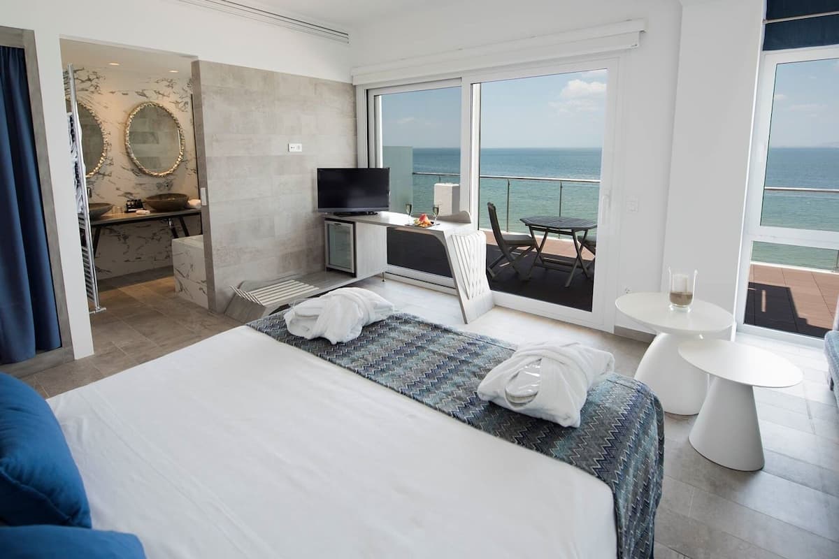 Una habitación de hotel moderna con una cama grande, un escritorio con televisor, un balcón con mesa y sillas, y vistas al mar.