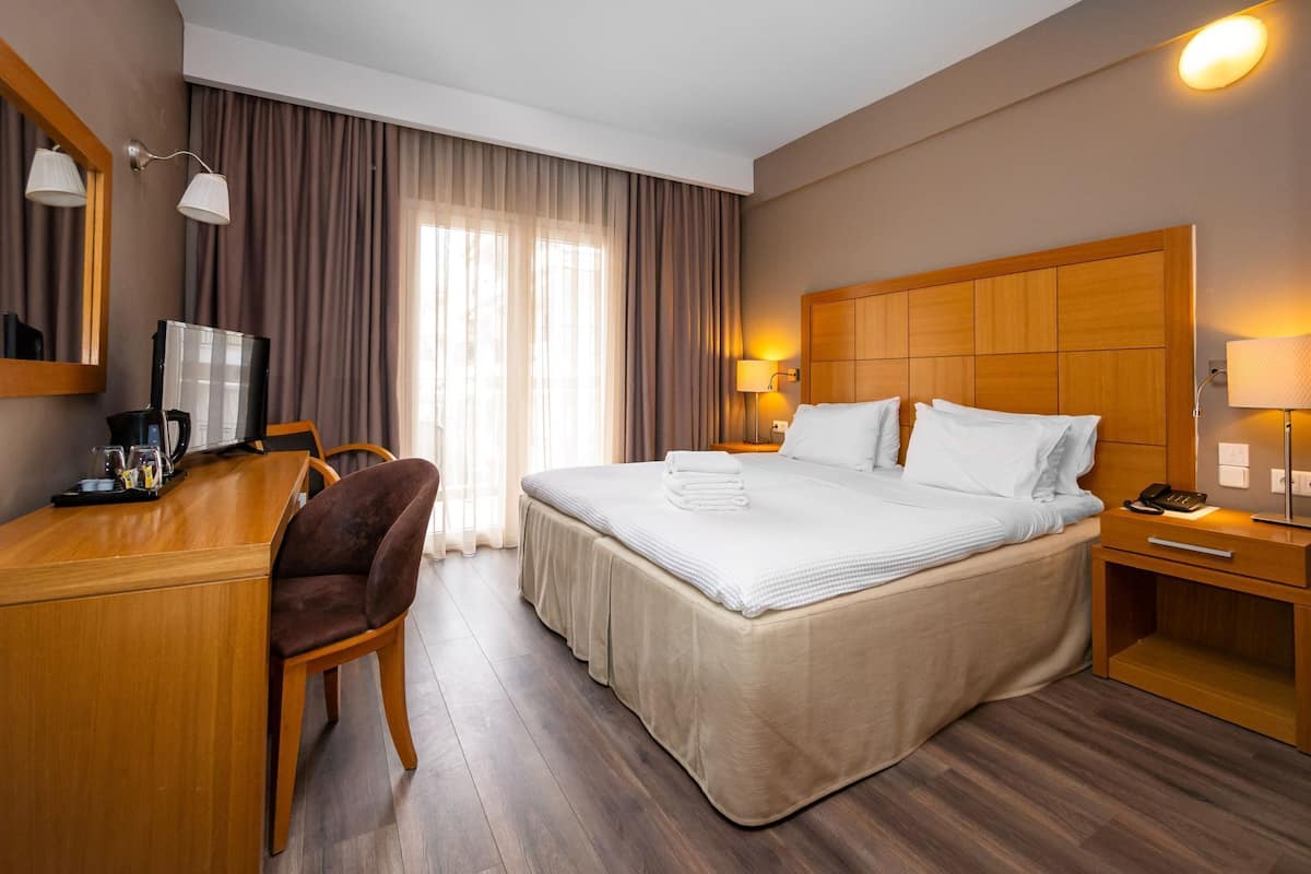 A hotel room with a bed, a desk with a coffee maker, a chair, and a nightstand with a lamp.
