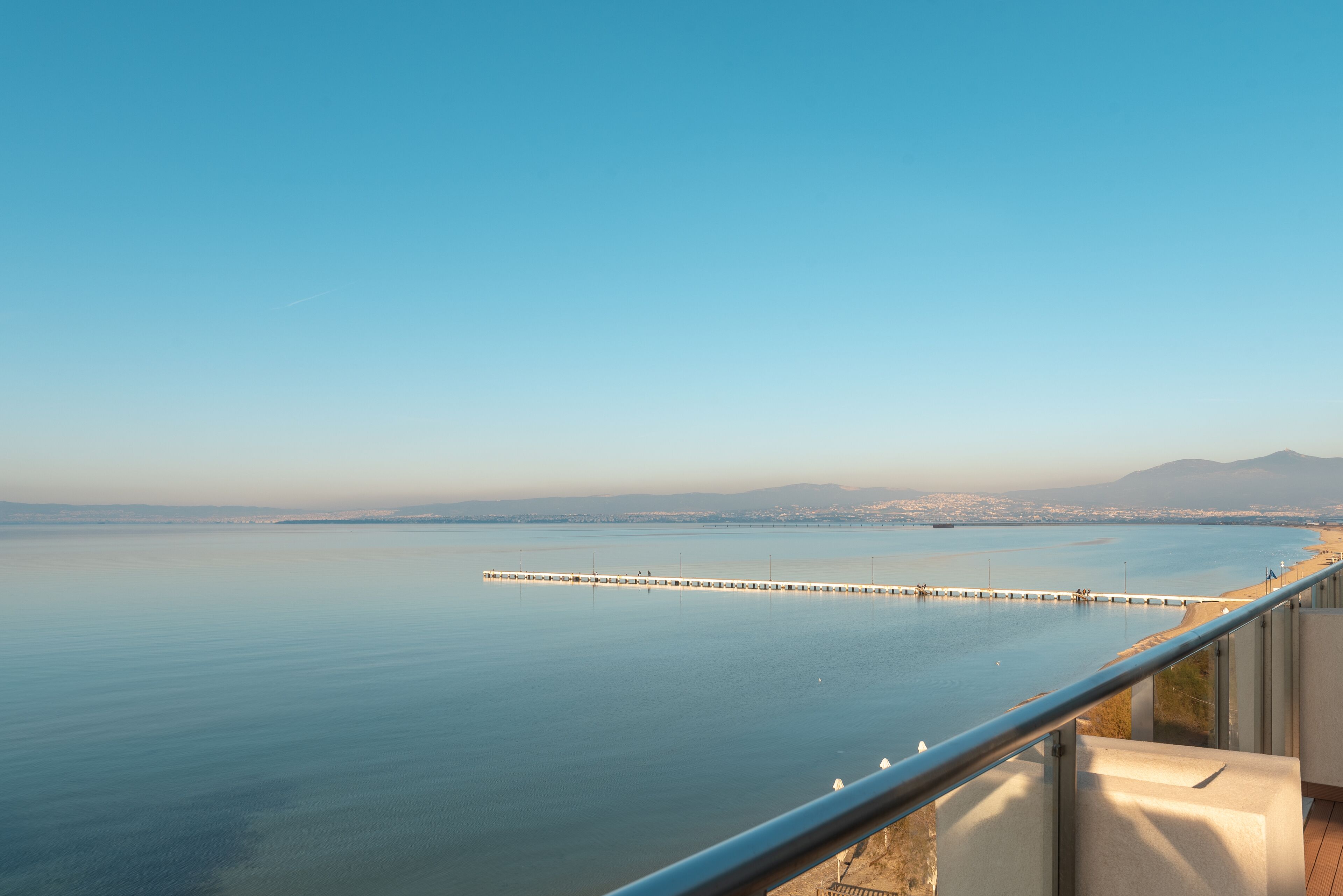Vista costanera des d'un balcó, amb cel serè, mar tranquil·la i un llarg moll que s'endinsa a l'aigua.