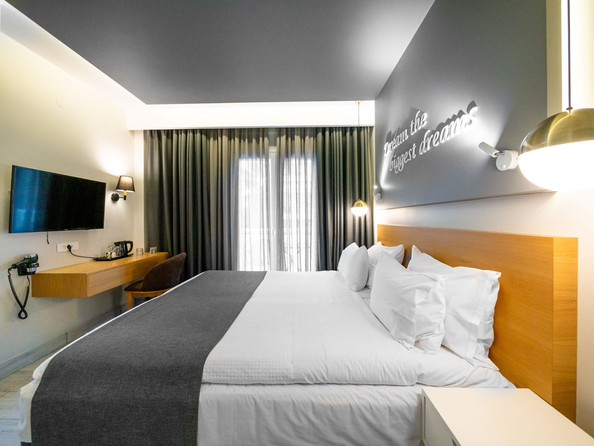 Habitación de hotel con cama, televisión, escritorio con lámpara y ventana con cortinas.