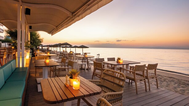 Un área de comedor al aire libre con mesas de madera y sillas de mimbre, con vista a una playa al atardecer.