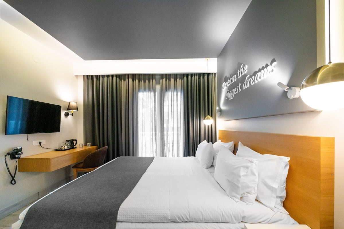 Habitación de hotel con una cama grande, un televisor de pantalla plana montado en la pared, un escritorio con una lámpara y una ventana con cortinas.