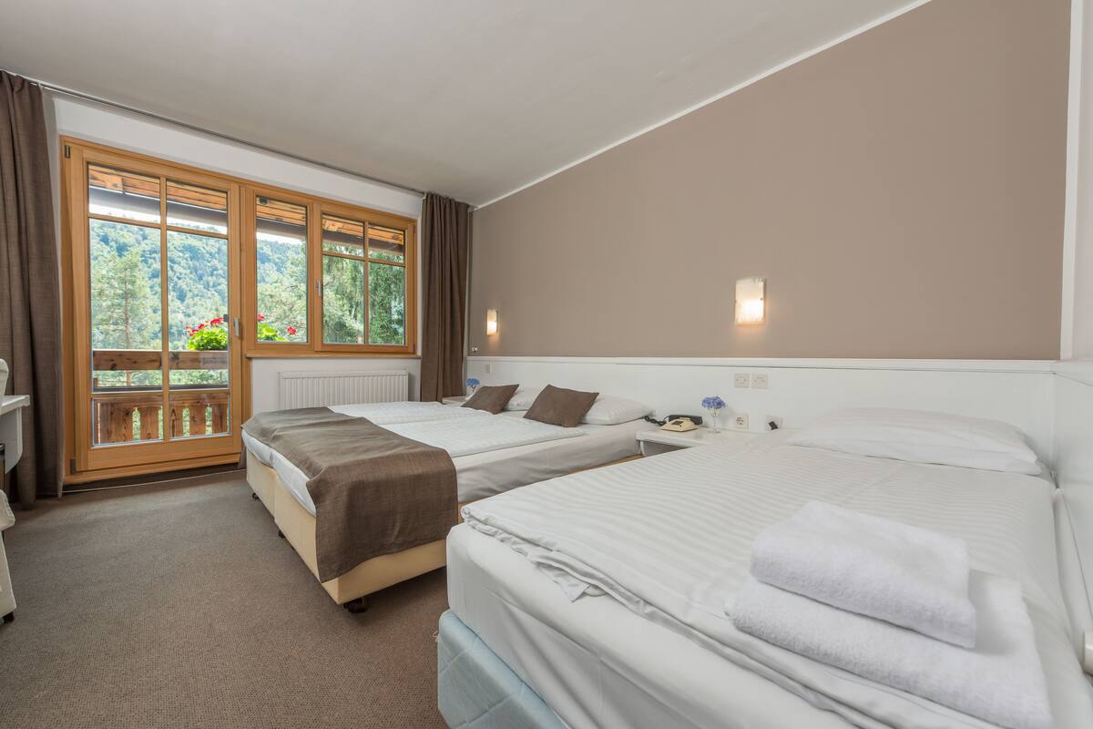 A hotel room with two beds, a window with a view of greenery, and a small table with a lamp.