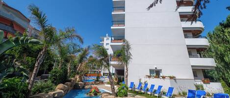 A hotel with a swimming pool, blue lounge chairs, and a palm tree.