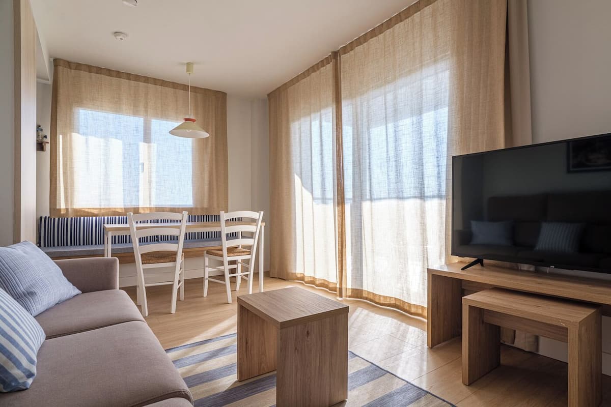 Ein modernes Wohnzimmer mit einer Couch, einem Fernseher, einem Couchtisch aus Holz, einem Esstisch mit Stühlen und großen Fenstern mit Vorhängen.