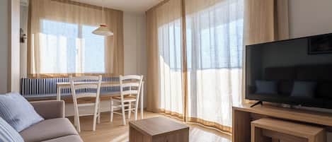 A modern living room with a sofa, a TV, a wooden coffee table, a dining table with chairs, and large windows with curtains.
