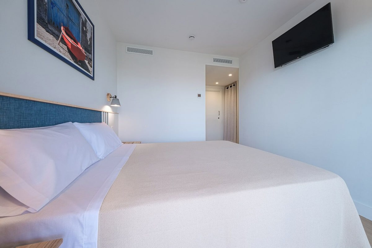 Ein Hotelzimmer mit Bett, einem an der Wand befestigten Fernseher, einem Gemälde und einer Tür zu einem weiteren Raum.