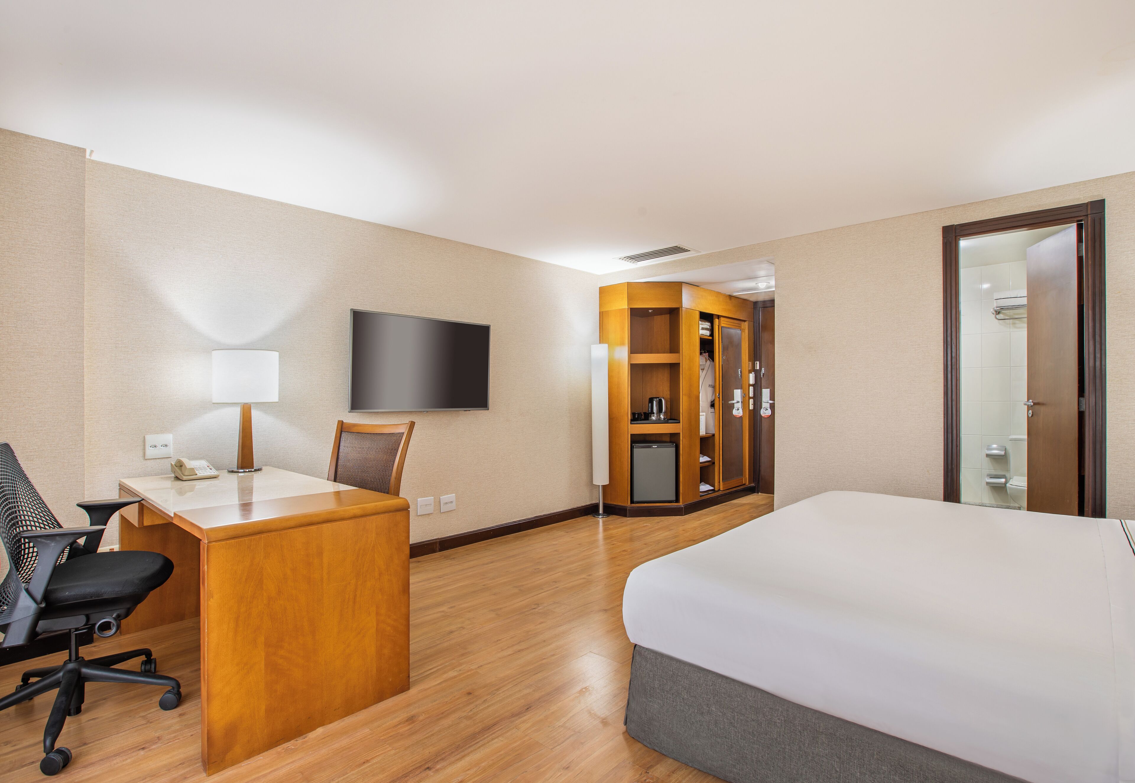 A hotel room with a wooden desk, a chair, a bed, a television, and a wardrobe.