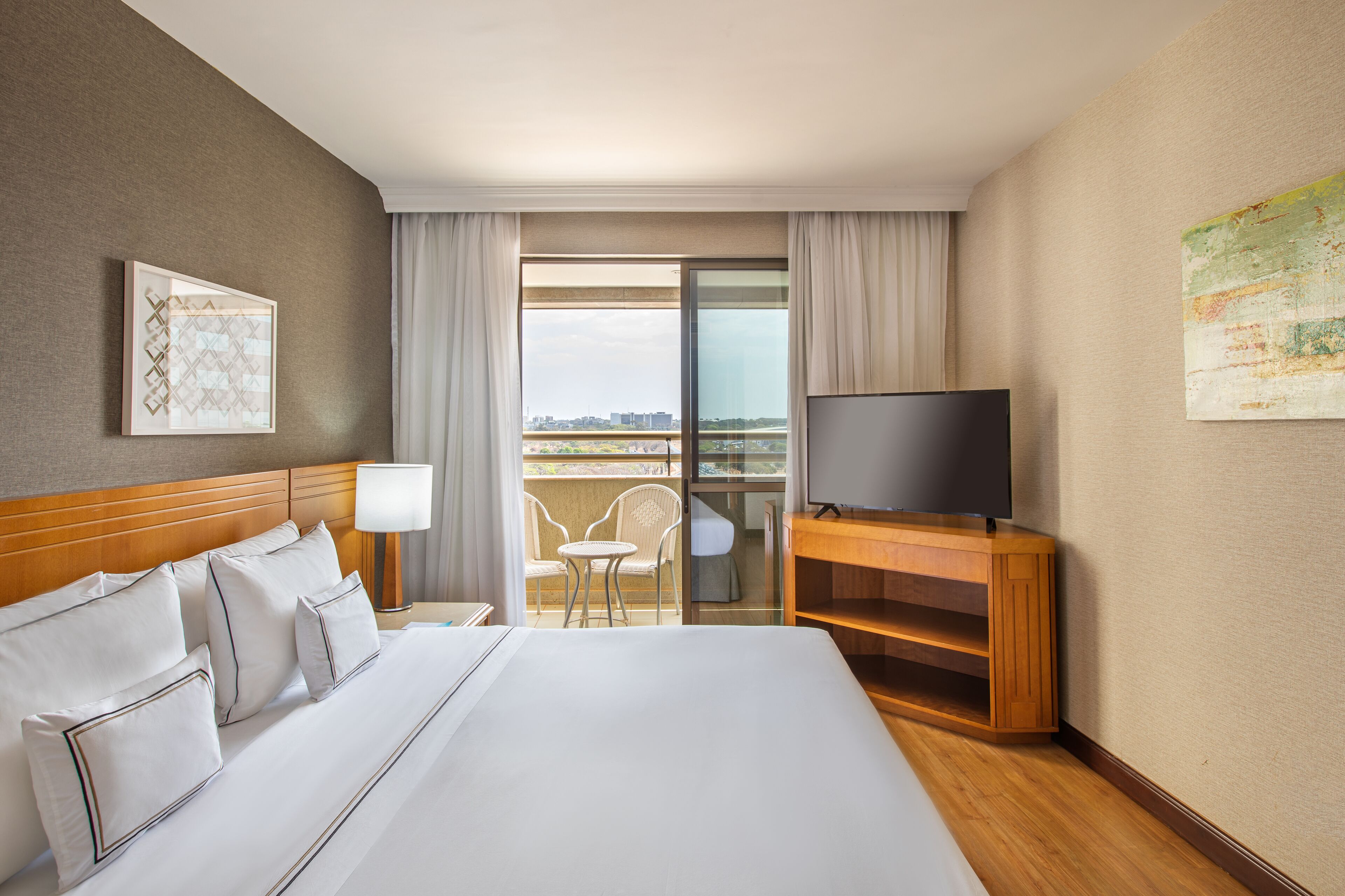 A hotel room with a large bed, a TV on a wooden stand, a balcony with chairs, and a city view.