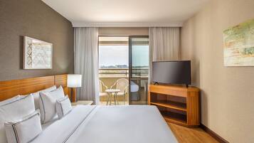 A hotel room with a large bed, a TV on a wooden stand, a balcony with chairs, and a city view.