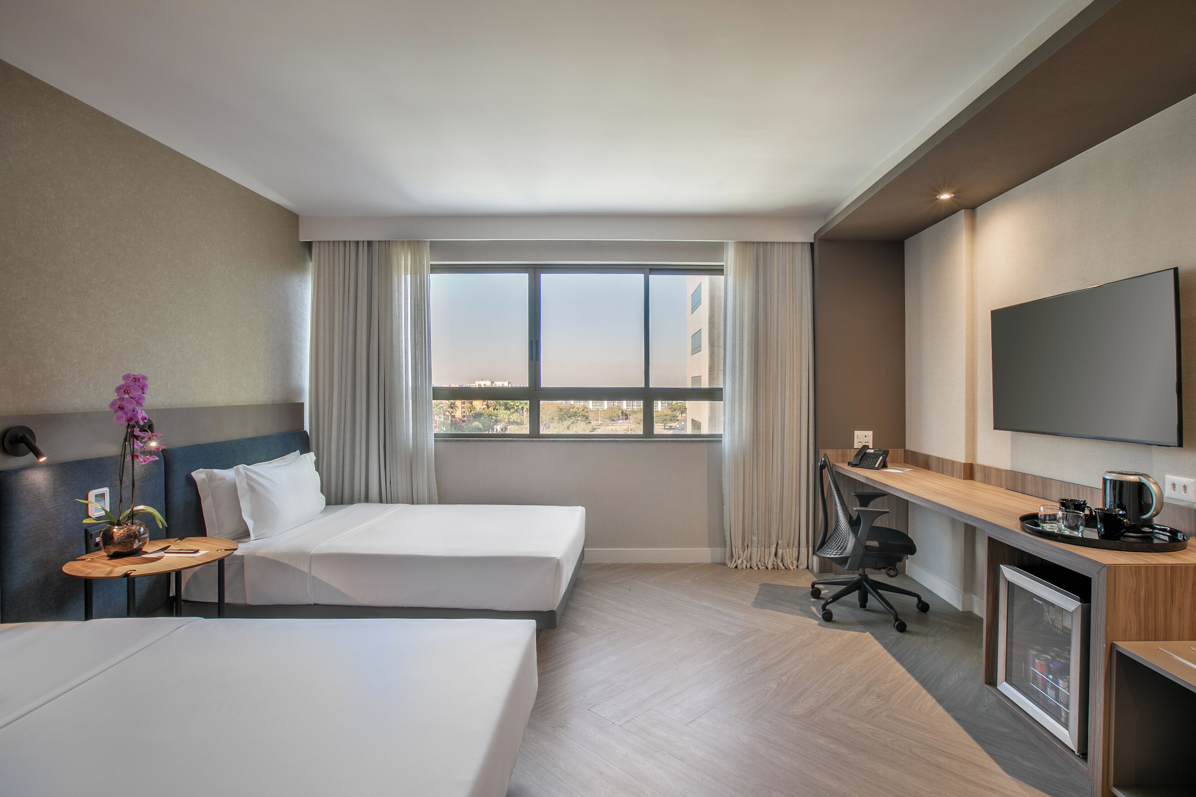 Habitación de hotel con dos camas, un escritorio, una silla, un televisor y ventana con vista a la ciudad.