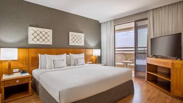 Une chambre d’hôtel avec un grand lit, une tête de lit en bois, deux tables de chevet avec des lampes, un téléviseur à écran plat et un balcon avec une porte vitrée.