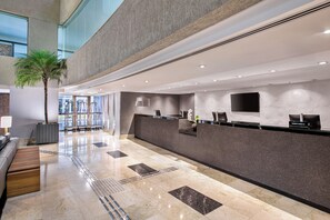 Een hotel receptie met een marmeren vloer, een receptiebalie, een palmboom en een groot raam.