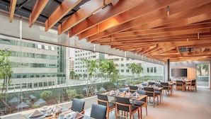 A modern dining area with tables set for service, large windows offering views of the city, and a high ceiling with wooden beams. - Melia Brasil 21 (Brasília)