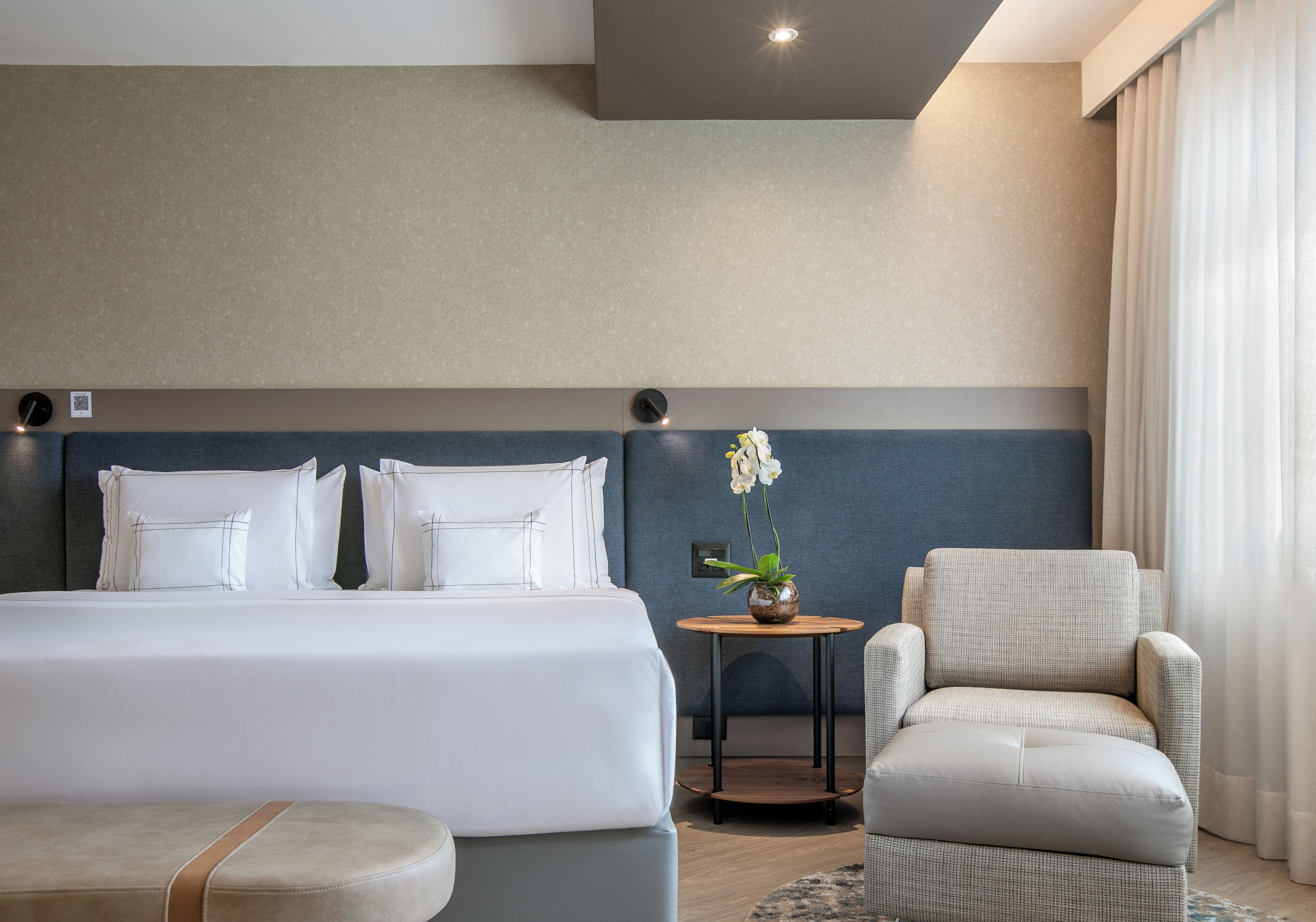 A hotel room with a bed, a chair, a small table with a plant, and a window with curtains.