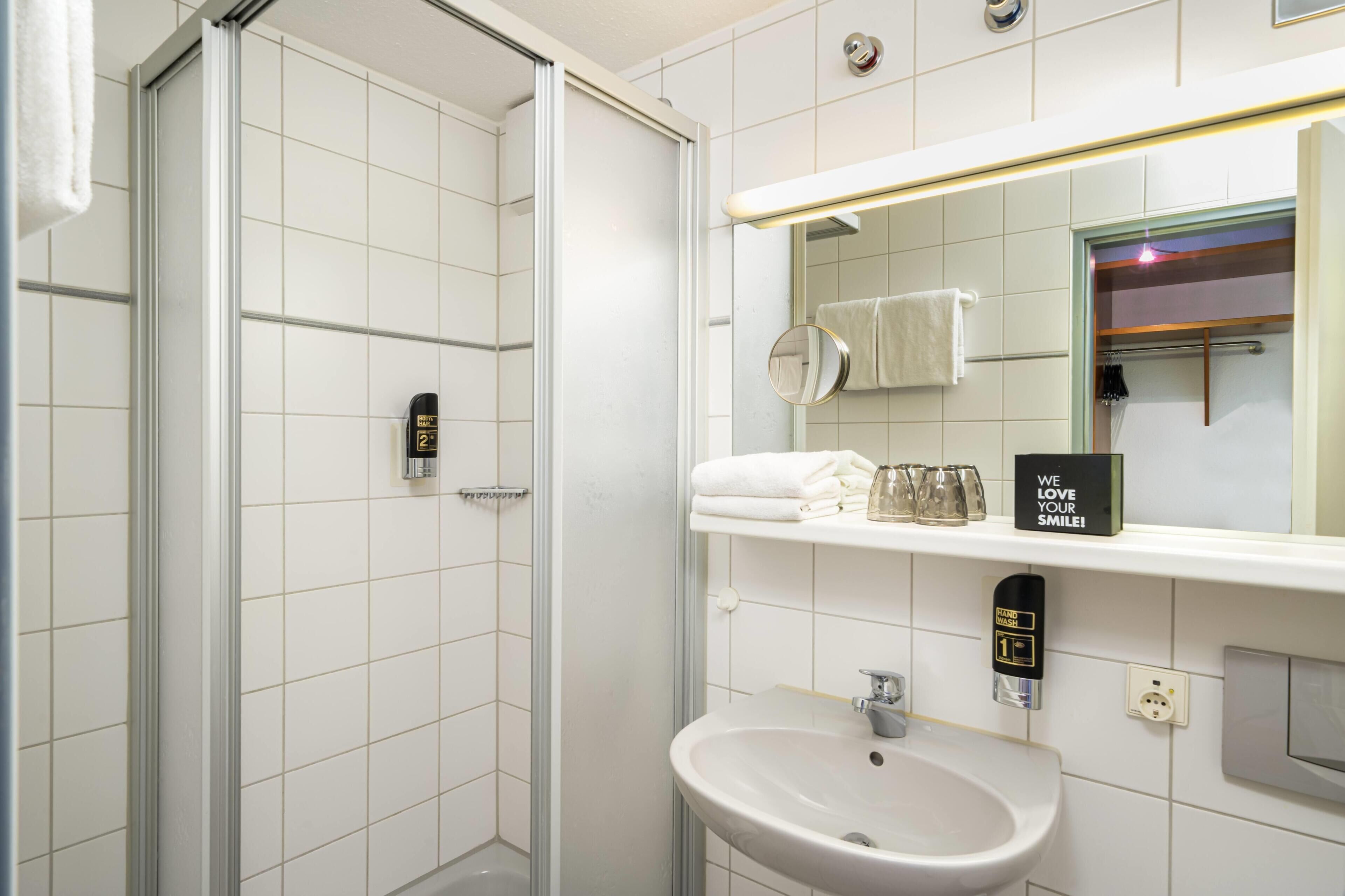 Un baño moderno con cabina de ducha de vidrio, un lavamanos, un espejo y una repisa con toallas y vasos.