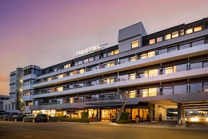 Un hotel de varios pisos con diseño moderno, que cuenta con balcones en cada planta y una entrada destacada con puertas de vidrio.