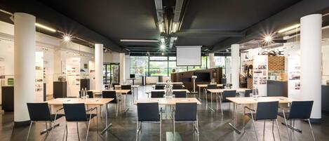 A spacious conference room with tables and chairs, large windows, and a modern design with exposed pipes and lighting fixtures.