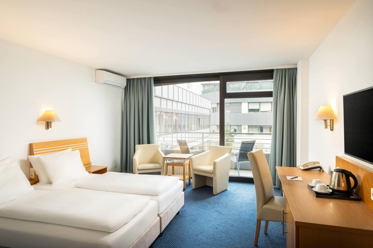 A hotel room with a bed, two chairs, a desk, a TV, and a window with curtains.