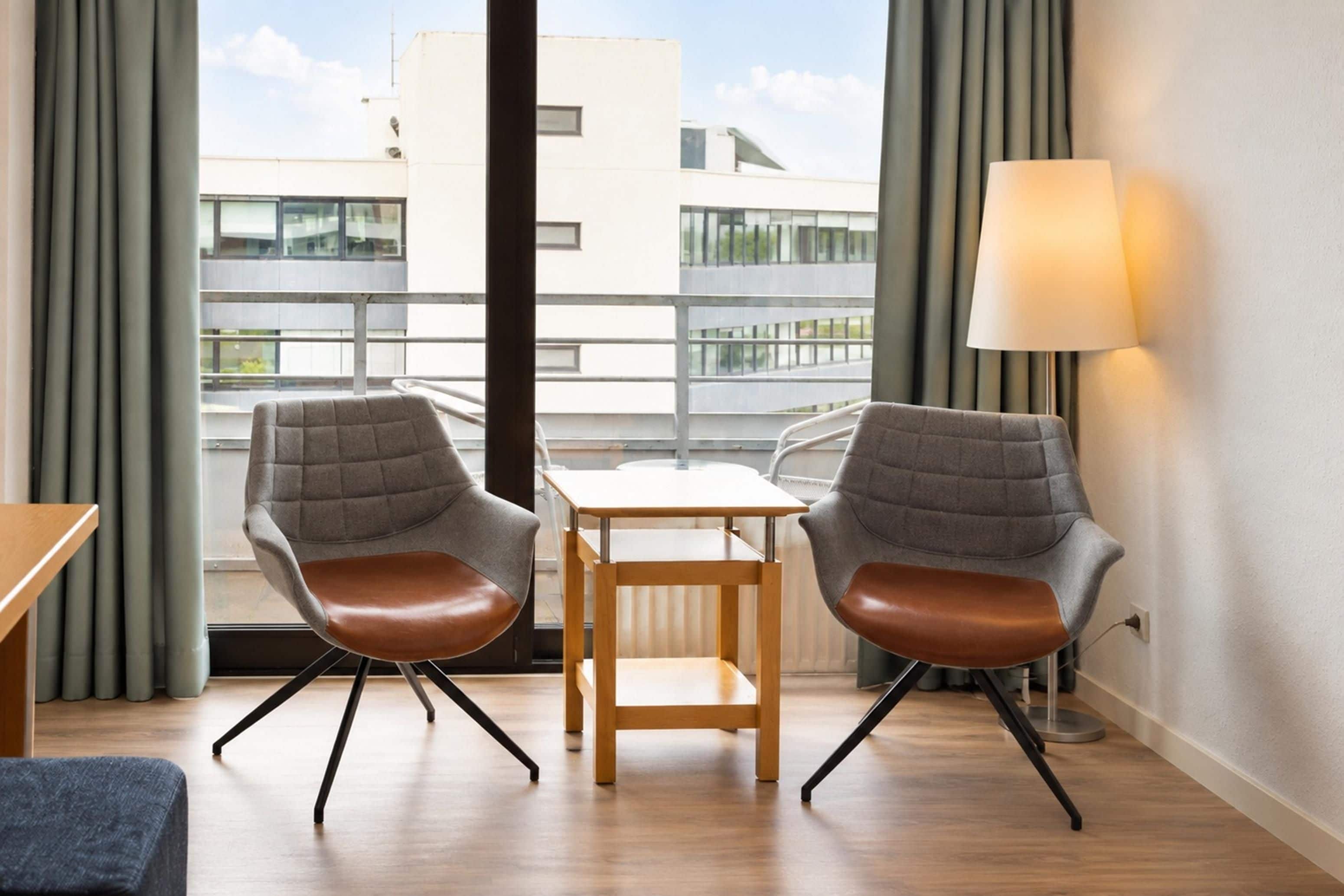 Ein modernes Hotelzimmer mit zwei grauen Polstersesseln, einem Holztisch, einer Stehlampe und Blick auf Gebäude durch große Fenster.