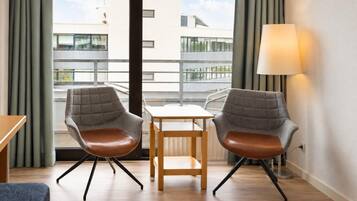 Ein modernes Hotelzimmer mit zwei grauen Polstersesseln, einem Holztisch, einer Stehlampe und Blick auf Gebäude durch große Fenster.