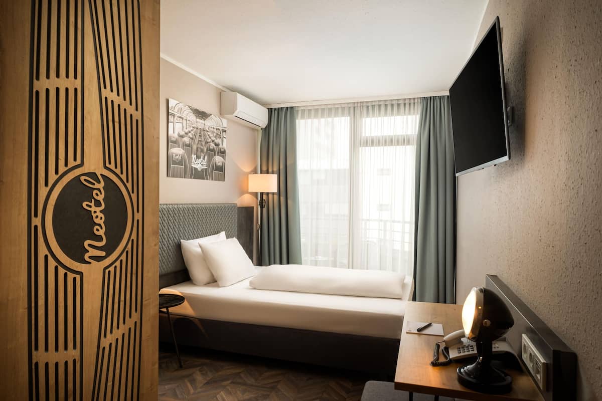 A hotel room with a bed, a desk with a telephone and lamp, a TV mounted on the wall, and a window with curtains.