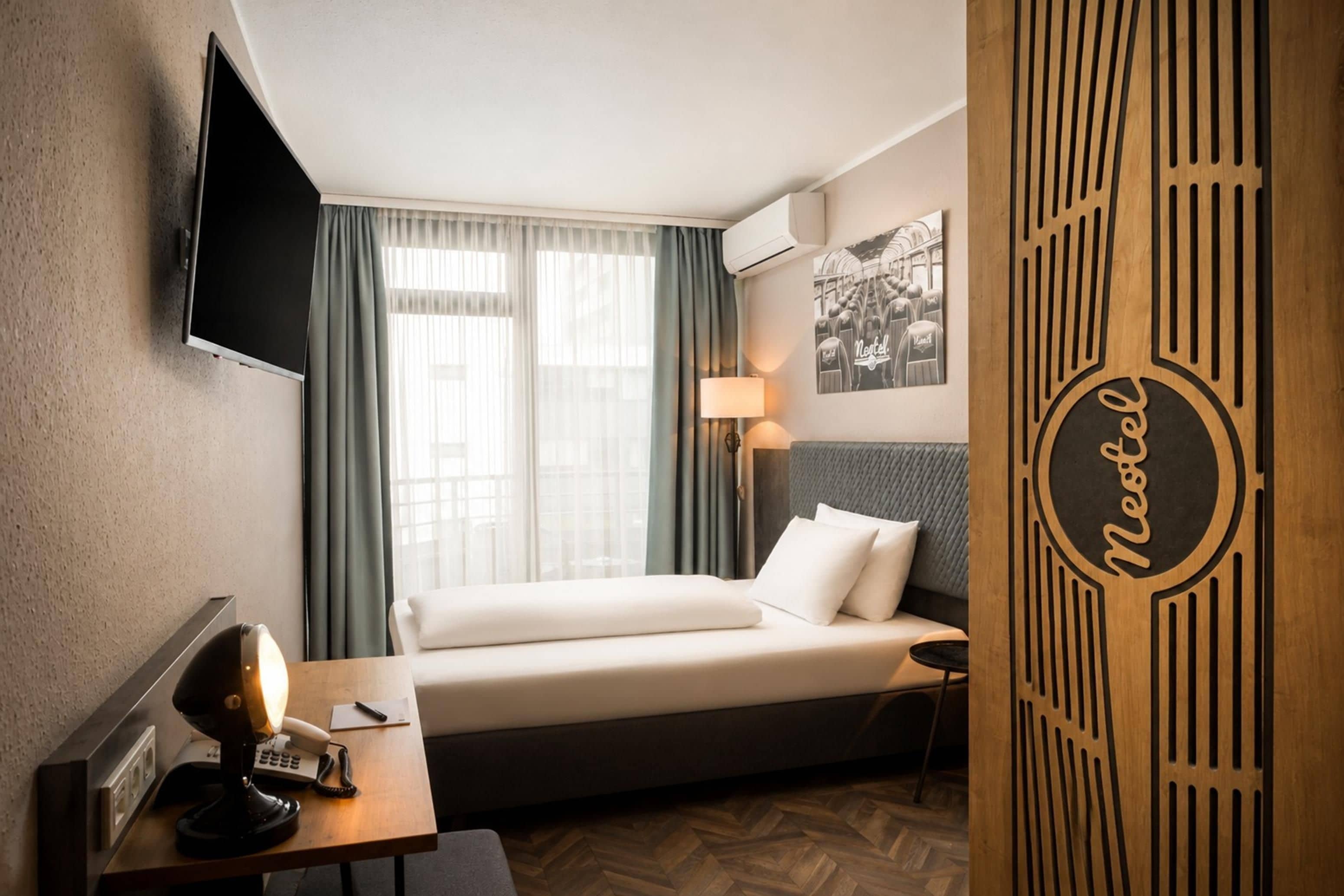 A hotel room with a bed, a desk with a telephone, a lamp, and a wall-mounted TV.