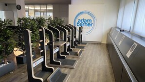 Un área de recepción con el logotipo de 'adam barner' en la pared, una fila de mostradores negros y blancos, y plantas en macetas.