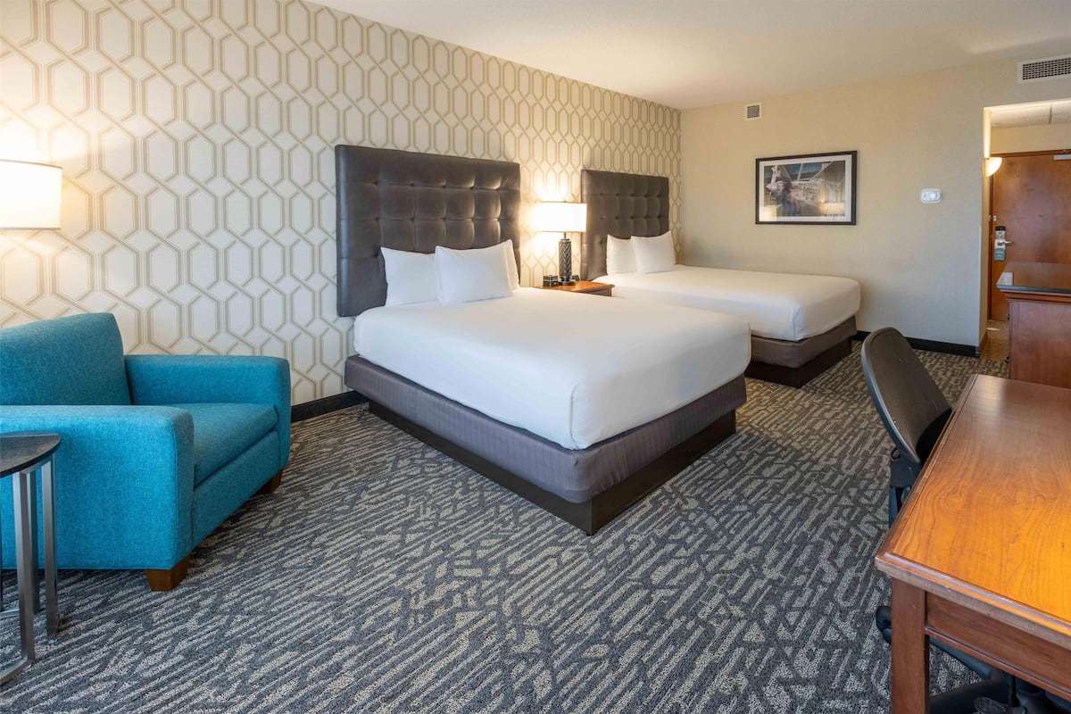 Una habitación de hotel con dos camas, un sillón azul, un escritorio de madera y una alfombra estampada.