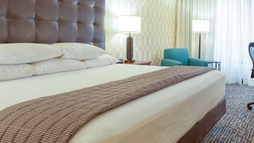 Habitación de hotel con una cama grande, cabecera, dos mesitas de noche con lámparas, un sillón azul y una ventana con cortinas.