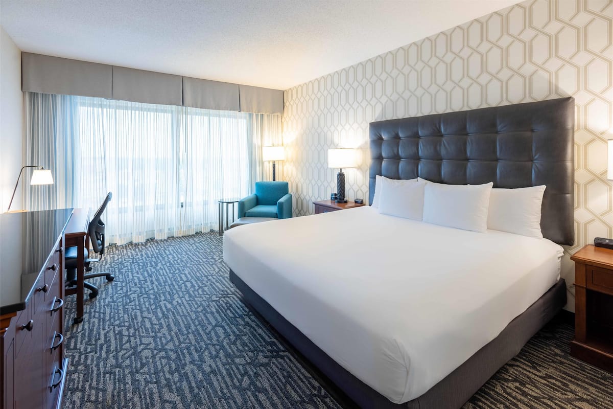 Habitación de hotel con una cama grande, un escritorio con cajones, una silla, dos lámparas de noche y suelo alfombrado.