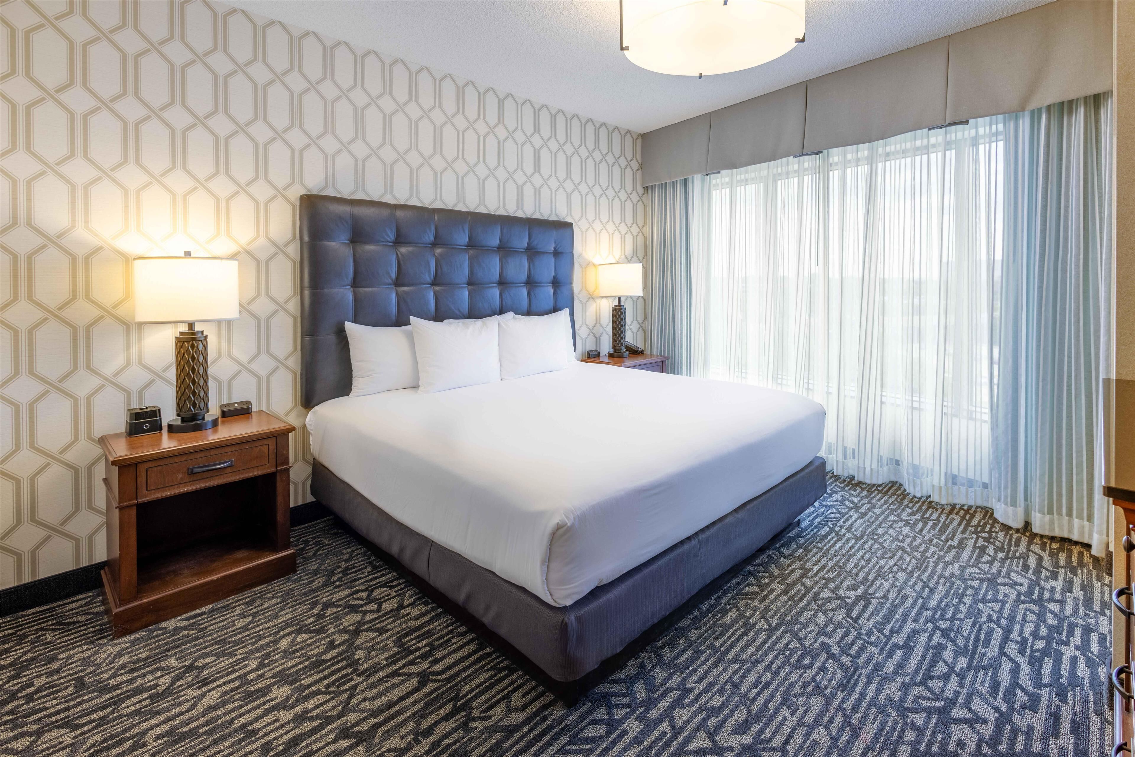 Quarto de hotel com uma cama grande, duas mesas de cabeceira com abajures, um tapete estampado e uma janela com cortinas finas.