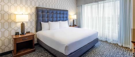 Quarto de hotel com uma cama grande, duas mesas de cabeceira com abajures, um tapete estampado e uma janela com cortinas finas.
