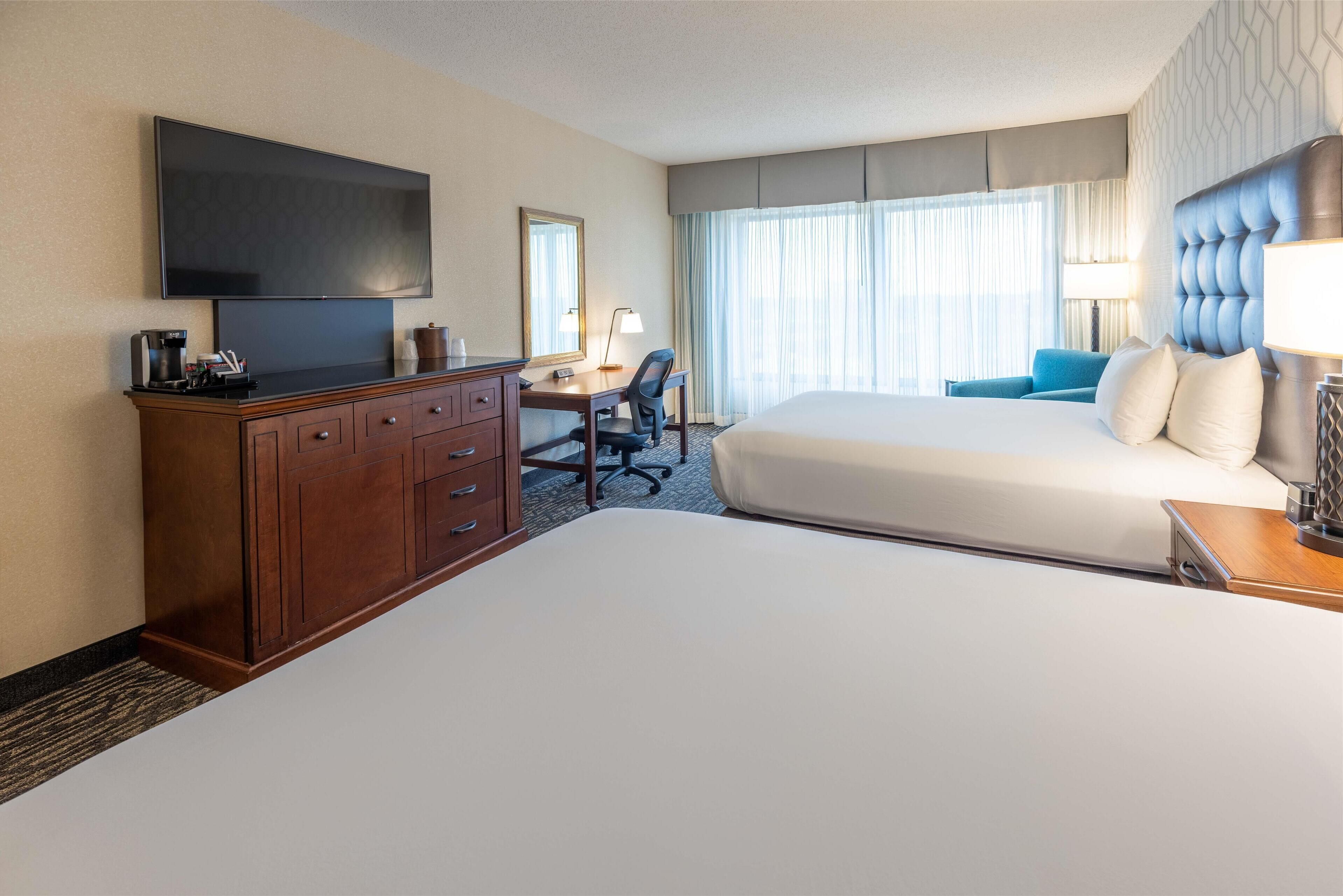 A hotel room with a large bed, a smaller bed, a desk with a chair, a television, and a window with curtains.